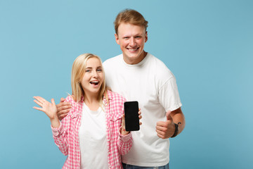 Funny couple two friends guy girl in white pink t-shirts posing isolated on blue background. People lifestyle concept. Mock up copy space. Hold mobile phone with empty blank screen, showing thumb up.