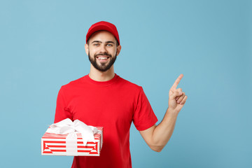 Delivery man in red uniform hold present box isolated on blue wall background, studio portrait. Professional male employee in cap t-shirt print working as courier. Service concept. Mock up copy space.