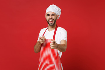 Bearded male chef cook or baker man in striped apron t-shirt toque chefs hat posing isolated on red background. Cooking food concept. Mock up copy space. Blinking, pointing index fingers on camera.