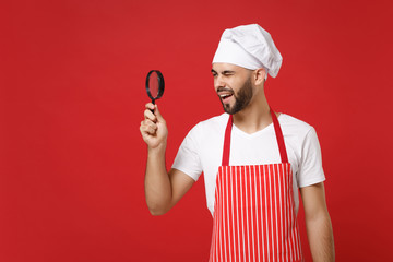 Curious young bearded male chef cook or baker man in striped apron white t-shirt toque chefs hat isolated on red wall background. Cooking food concept. Mock up copy space. Holding magnifying glass.