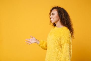 Side view of smiling young african american girl in fur sweater posing isolated on yellow orange background. People lifestyle concept. Mock up copy space. Standing with outstretched hand for greeting.