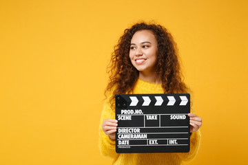 Smiling young african american girl in fur sweater posing isolated on yellow orange background in studio. People lifestyle concept. Mock up copy space. Holding classic black film making clapperboard. - Powered by Adobe