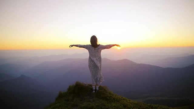 Young beautiful woman in a long dress developing in the wind at the top of the mountain at sunset