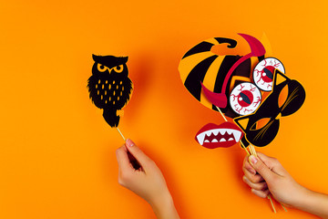 Female hands are holding funny paper photo props on orange background. Black owl, cat mask, horns, vampire teeth, blood eyes, witch hat on canvas. Party accessories for celebrating happy halloween.