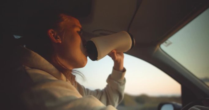 Young blond man drinks from water bottle while driving in the light of the setting sun in the car