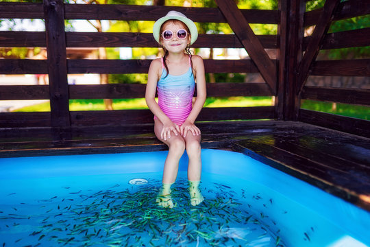 Young girl in hat, swimsuit receiving massage with small fishes. Peeling with fish. Girl enjoying medicinal procedure. Foot massage with fish in aquarium closeup. Fish spa procedure.