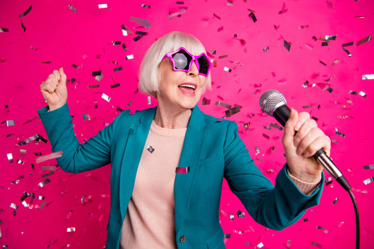 Photo of old mature stylish energetic woman singing in microphone wearing star shaped spectacles standing in falling confetti isolated over pink vivid color background