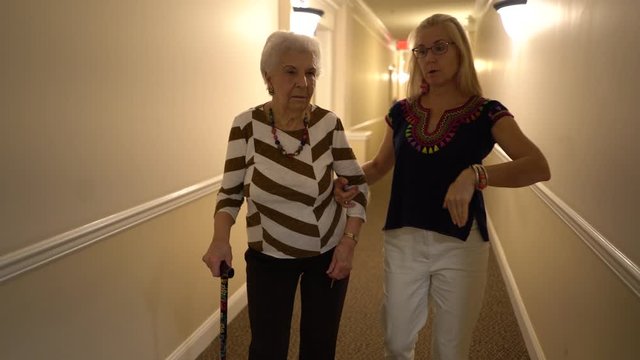 Pretty woman in ethnic blouse holds elderly woman&rsquo;s arm and they walk down a hallway.