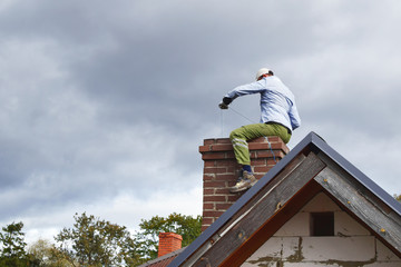 Chimney sweep man cleaning brown brick chimney while sitting on chimney on building roof on cloudy...