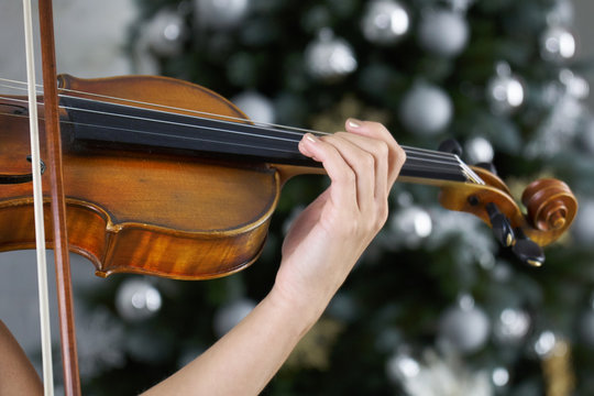 Hand of a female violinist on the fingerboard of a violin takes a chord with a decorated Christmas tree on the background.Classical music concert on Christmas or New Year eve