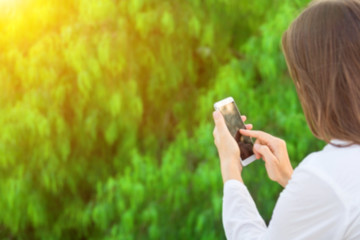 Young caucasian woman holds white smartphone touching screen texting reading on green foliage background in sunlight. Modern technology communication concept. Blurred defocused template