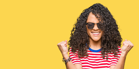 Young beautiful woman with curly hair wearing sunglasses celebrating surprised and amazed for success with arms raised and open eyes. Winner concept.