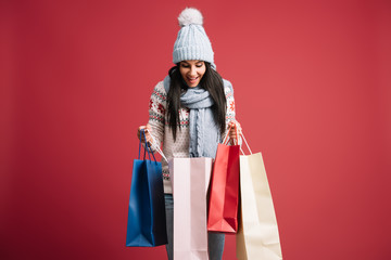 happy woman in winter sweater, scarf and hat looking into shopping bags, isolated on red