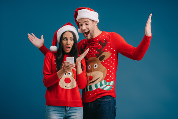 Obraz premium excited couple in santa hats and christmas sweaters using smartphone isolated on blue