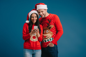 couple in santa hats and winter sweaters using smartphone isolated on blue