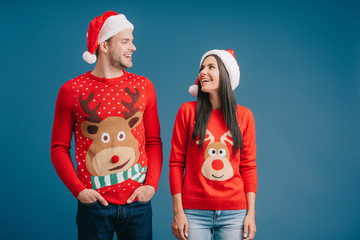 laughing couple posing in santa hats and sweaters isolated on blue