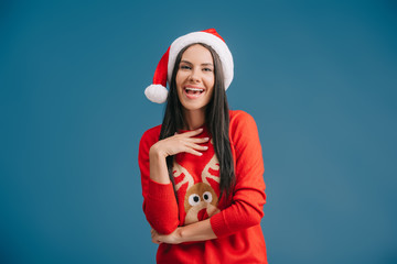 happy woman posing in santa hat and christmas sweater isolated on blue