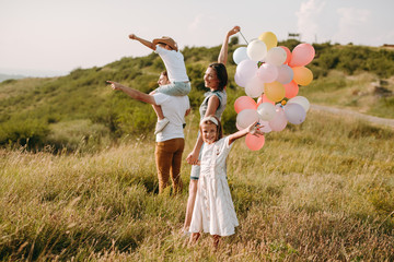 Beautiful family of four taking a walk in the green mountain fields carrying colourful baloons smiling and enjoying their holiday .