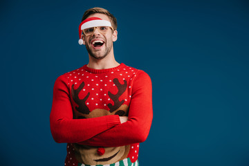 excited man posing in christmas sweater and eyeglasses with santa hat, isolated on blue