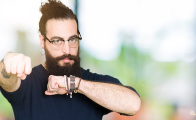 Young hipster man with long hair and beard wearing glasses Punching fist to fight, aggressive and angry attack, threat and violence