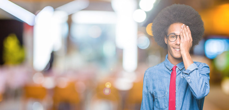 Young african american business man with afro hair wearing glasses and red tie covering one eye with hand with confident smile on face and surprise emotion.