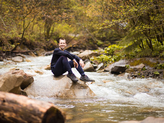 Traveler man wearing in black sitting on rock in mountain river between forest. Travel lifestyle concept
