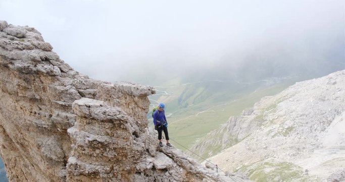 Woman on a ridge in Dolomites, Italy, with fog behind her in the valley, during an easy section of via ferrata Cesare Piazzetta route to Piz Boe peak - Summer adventure activities concept.