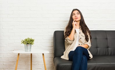 Young arab woman sitting on the sofa looking sideways with doubtful and skeptical expression.