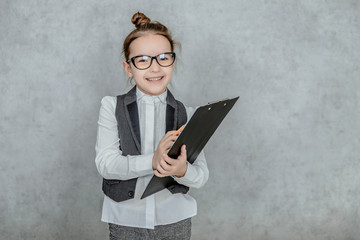 The little lady boss is on a gray background. During this, holds a black folder and a yellow pencil looking into the camera.