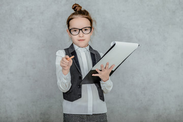 Little girl with a folder in her hands on a gray background. Dressed up as a business lady and glasses. Looking at the camera, bewildered, showing a pencil to the camera.