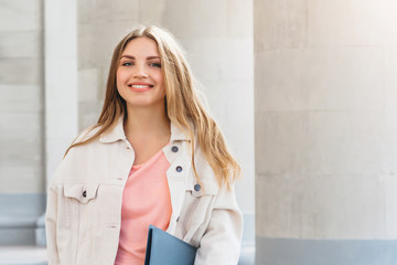 Young blonde girl student smiling against university. Cute girl student holds folders and notebooks in hands, copy space. Learning, education concept