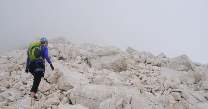 Female hiker equipped with climbing gear ascending on a path through big rocks, towards fog and low clouds, at the end of the via ferrata route Cesare Piazzetta to Piz Boe.