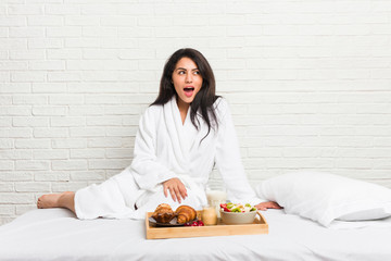 Young curvy woman taking a breakfast on the bed being shocked because of something she has seen.