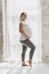 Long shot pregnant woman posing next to window
