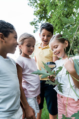 happy multicultural kids looking at green leaves though magnifier