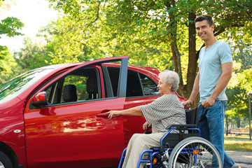 Young man helping disabled senior woman in wheelchair to get into car outdoors