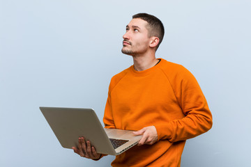 Young caucasian man holding a laptop smiling confident with crossed arms.