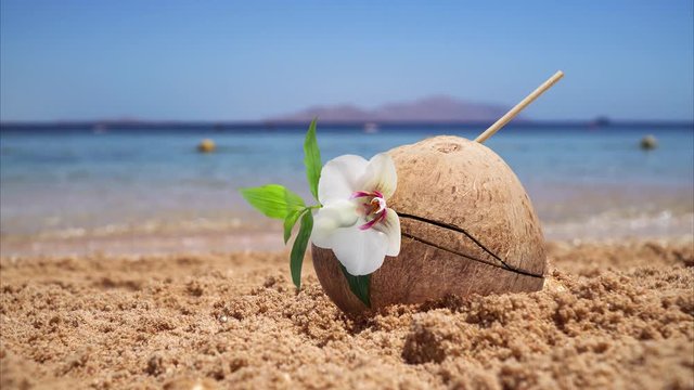 Coco cocktail in a coconut stands in the sand on an ocean beach. Small waves rise behind. Female legs in defocus pass by.