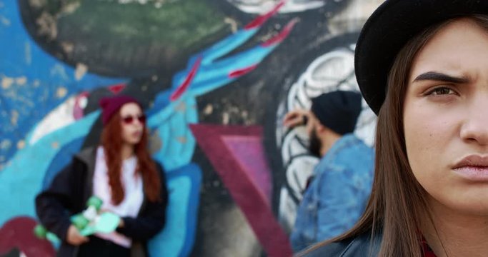 Close up of the half face of young beautiful hipster woman in black hat looking straight with sad and worried face like being angry. Couple on the background at the wall with graffity. Love triangle.