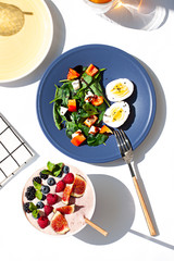 Healthy sunny morning breakfast on the white table. A Smoothie bowl and A Salad with spinach, red pepper, feta cheese eggs with boiled eggs. Top view