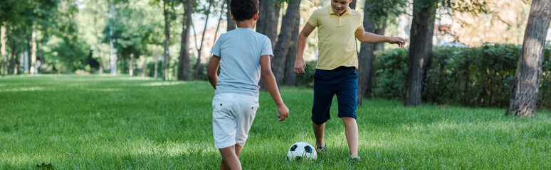 panoramic shot of cute multicultural boys playing football on green grass