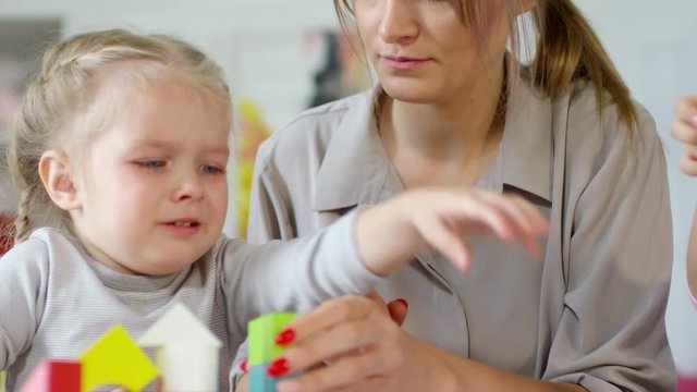 Upset little girl with tears on her face building toy block tower with help of female teacher while playing in kindergarten