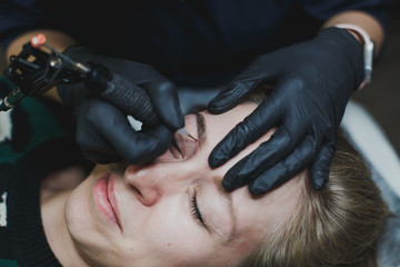 master in black gloves does permanent eyebrow makeup to a young beautiful blonde girl 1