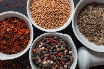 Paprika, mustard, caraway seeds and a mixture of peppercorns in white ceramic bowls next to the mortar isolated on black background