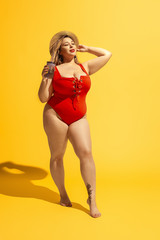 Young caucasian plus size female model's preparing for beach resort on yellow background. Woman in red swimsuit and hat drinking cocktail. Concept of summertime, party, body positive, equality.