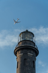 Fototapeta na wymiar Flying passenger jet airplane passes over lighthouse monument on clear summer day past blue sky and clouds. Light houses used for aviation and maritime navigation with circular strobe spot light