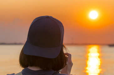 Female photographer takes picutres of a beautiful orange sunset over the water wearing backwards blue hat. Over the shoulder composition.