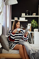 Beautiful smiling caucasian brunette dressed in sweater sitting on bed in bedroom, using tablet and drinking coffee in morning.