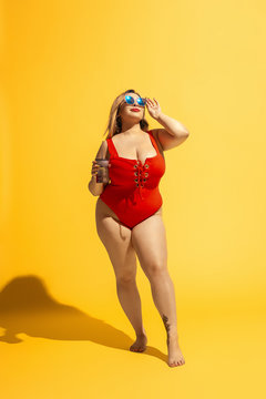 Young caucasian plus size female model's preparing for beach resort on yellow background. Woman in red swimsuit, hat and sunglasses drinking cocktail. Concept of summertime, party, body positive.