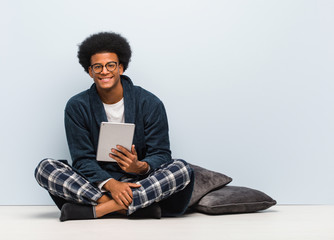 Young black man sitting on his house and holding his tablet cheerful with a big smile